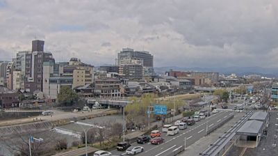 Kyoto daytime — Kamo River with exposed stone riverbed, traffic on bridge, modern and traditional buildings, mountains visible through low skyline