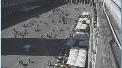Piazza San Marco from above — people scattered across the vast square, cafe tables along the arcade, self-organizing crowd patterns